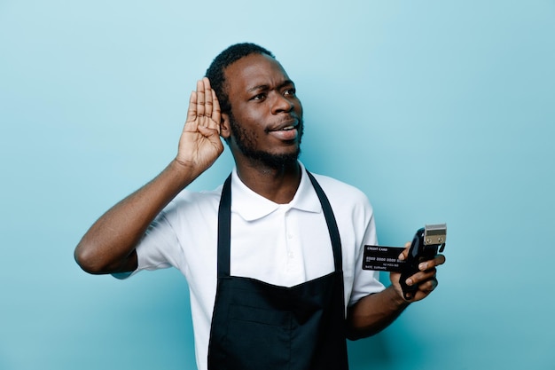 Глядя на сторону, показывающую слушающий жест, держащий карточку с машинкой для стрижки волос, молодой африканский парикмахер в униформе, изолированный на синем фоне