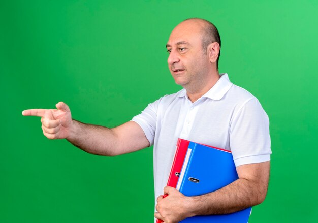 Looking at side smiling casual mature man holding folders and points at side isolated on green background