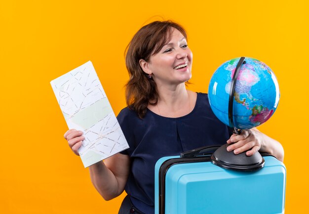 Looking at side smiling middle-aged traveler woman holding suitcase and globe with map on isolated yellow wall