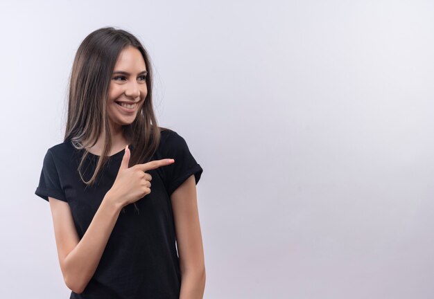 Глядя в сторону, улыбающаяся молодая кавказская девушка в черной футболке указывает в сторону пальцем на изолированной белой стене