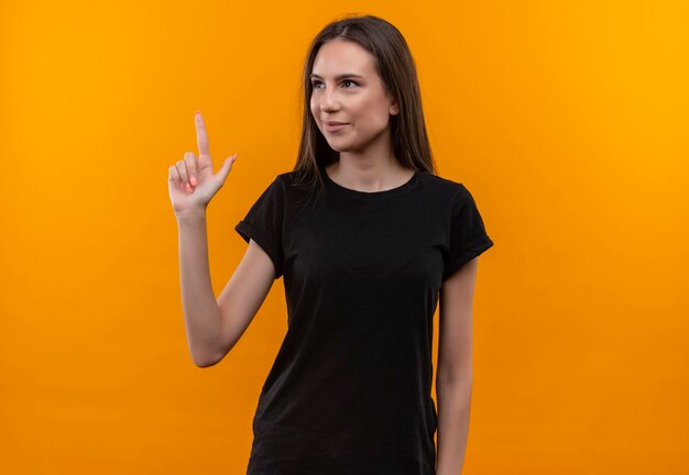 Глядя в сторону, улыбающаяся молодая кавказская девушка в черной футболке указывает пальцем на изолированную оранжевую стену