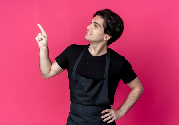 Looking at side smiling young handsome male barber in uniform points at side and putting hand on hip isolated on pink wall with copy space