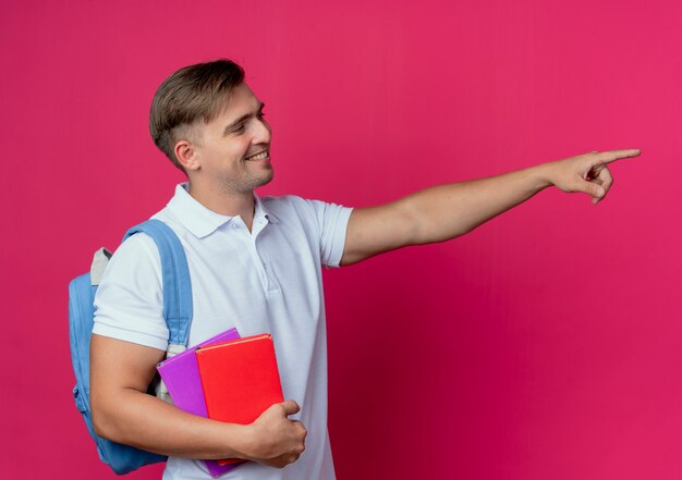 Looking at side smiling young handsome male student wearing back bag holding books and points at side