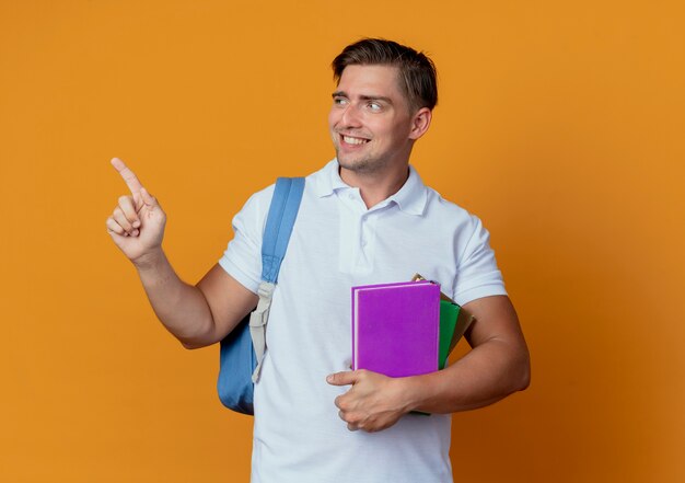 Looking at side smiling young handsome male student wearing back bag holding books and points at side
