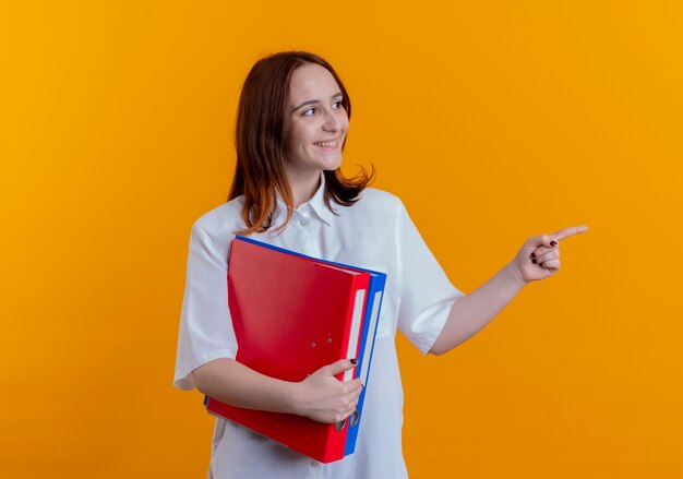 Looking at side smiling young redhead girl holding folders and points at side isolated on yellow background with copy space