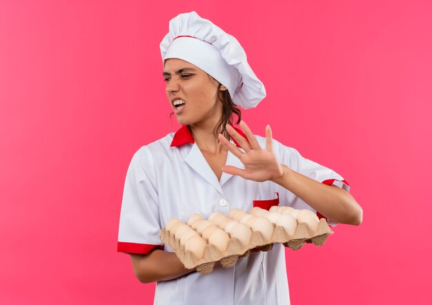 Глядя на сбоку, брезгливая молодая женщина-повар в униформе шеф-повара держит партию яиц на изолированной розовой стене с копией пространства