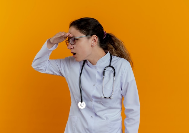 Глядя на сторону удивленной молодой женщины-врача в медицинском халате и стетоскопе в очках, глядя вдаль с рукой, изолированной на оранжевой стене