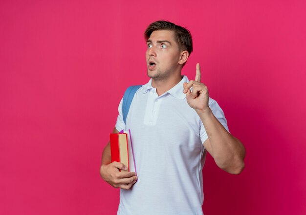 Looking at side surprised young handsome male student wearing back bag holding books and points at up isolated on pink wall