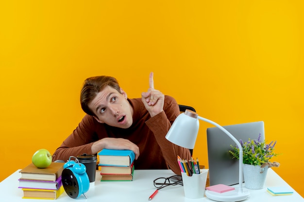 Looking at side surprised young student boy sitting at desk with school tools points at up  