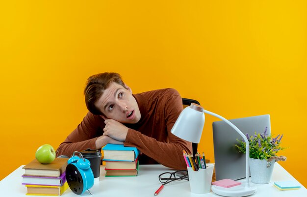 Looking at side surprised young student boy sitting at desk with school tools putting head on books 