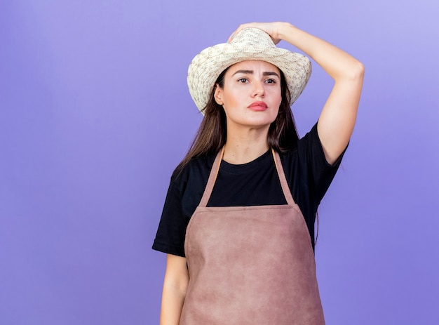 Free photo looking at side thinking beautiful gardener girl in uniform wearing gardening hat putting hand on hat isolated on blue background with copy space