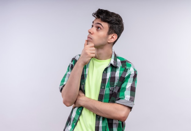 Looking at side thinking caucasian young man wearing green shirt put his hand on chin on isolated white wall