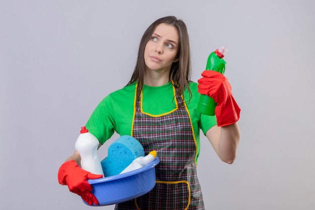 Looking at side thinking cleaning young woman wearing uniform in red gloves holding cleaning tools on isolated white wall