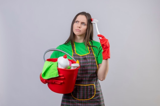 Looking at side thinking cleaning young woman wearing uniform in red gloves holding cleaning tools scratches head with cleaning brush on isolated white wall