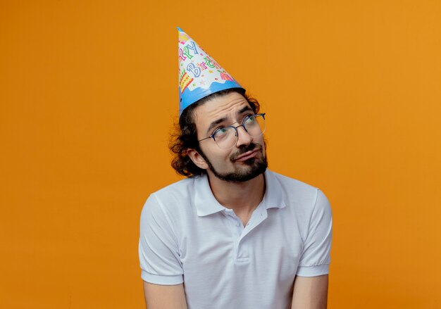 Looking at side thinking handsome man wearing glasses and birthday cap  isolated on orange background