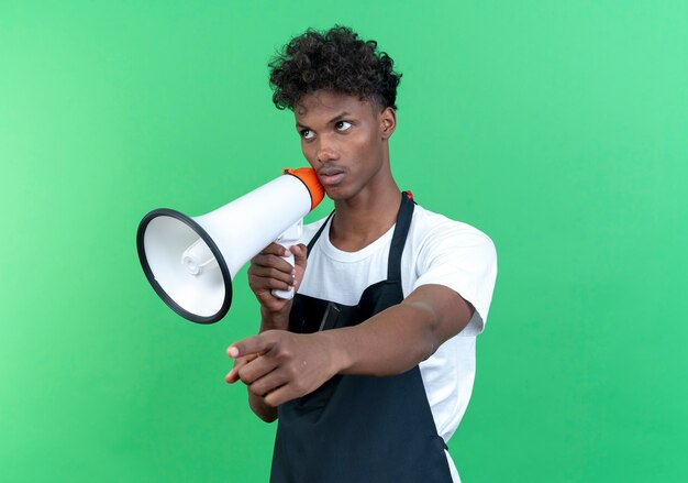 Looking at side thinking young afro-american male barber wearing uniform holding loudspeaker and points at side isolated on green background