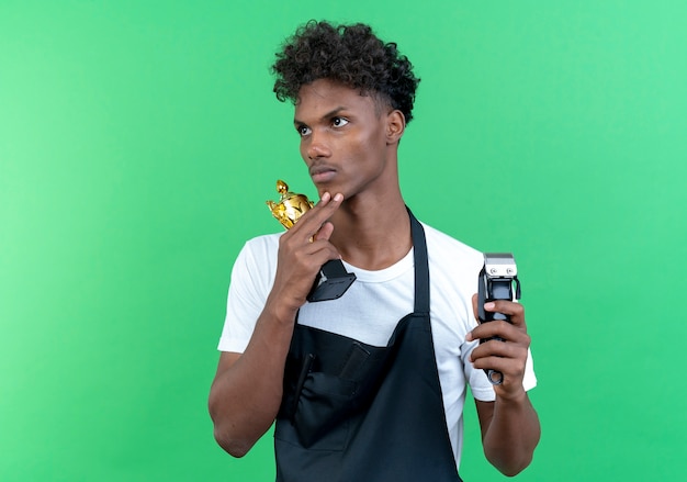 Looking at side thinking young afro-american male barber wearing uniform holding winner cup with hair clippers isolated on green background