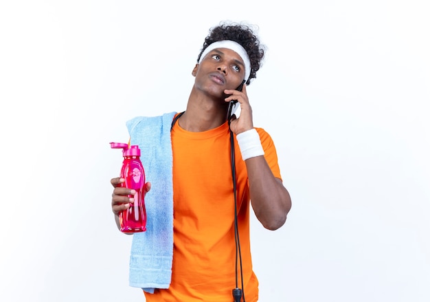 Looking at side thinking young afro-american sporty man wearing headband and wristband speaks on phone holding water bottle with towel and jump rope on shoulder