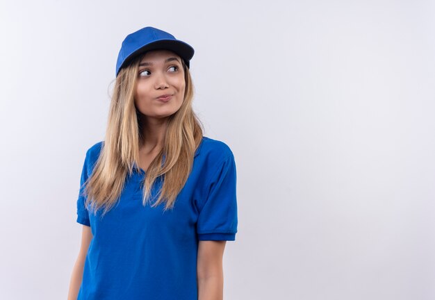 Looking at side thinking young delivery girl wearing blue uniform and cap isolated on white wall with copy space