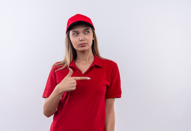 Глядя на сбоку, думая, что молодая доставщица в красной форме и кепке указывает в сторону, изолированную на белой стене с копией пространства