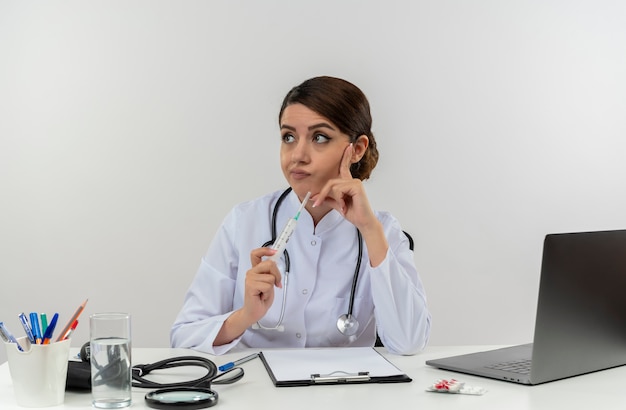 コピースペースのある孤立した白い壁に注射器を保持している医療ツールを備えたコンピューターのデスクワークに座って聴診器で医療ローブを着ている若い女性医師を考える側を見て