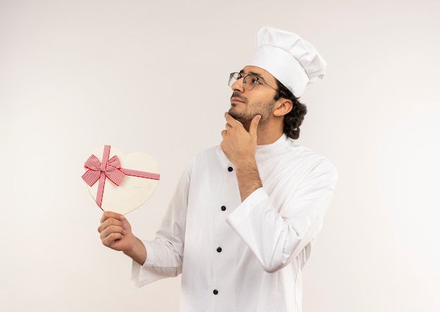Looking at side thinking young male cook wearing chef uniform and glasses holding heart shape gift box and putting hand on chin