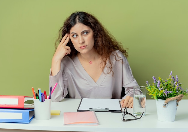 Глядя на сторону мышления молодой симпатичной женщины-офисного работника, сидящего за столом с офисными инструментами, положив палец на лоб, изолированные на оливковом фоне