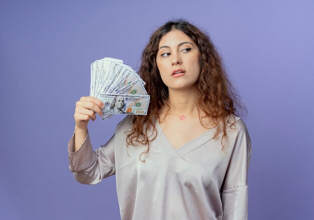 Looking at side thinking young pretty girl holding cash isolated on blue background