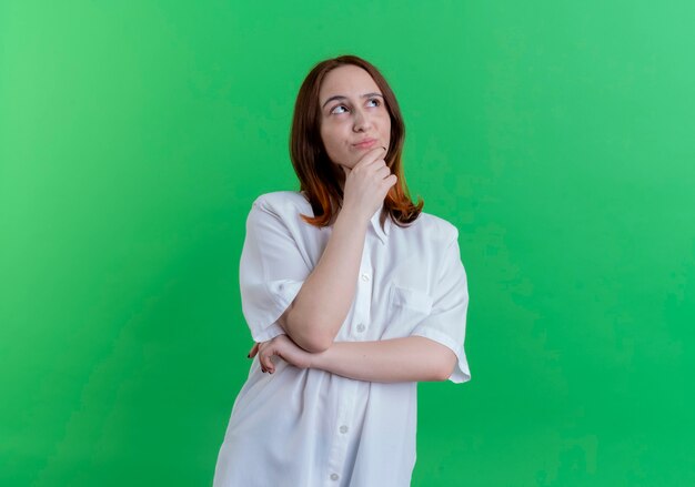 Looking at side thinking young redhead girl putting hand under chin isolated on green background with copy space