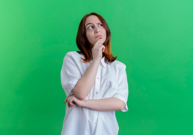 Looking at side thinking young redhead girl putting hand under chin isolated on green background