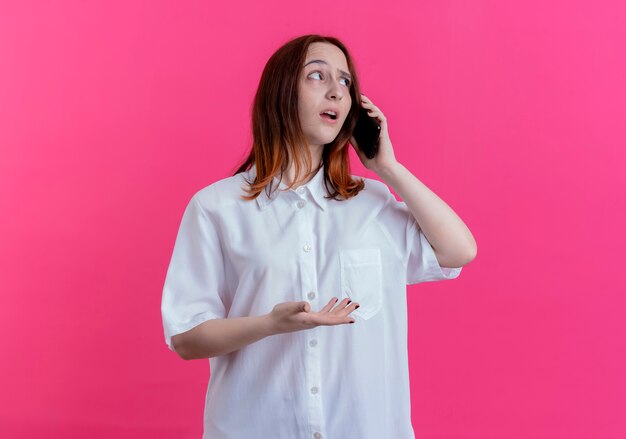 Looking at side thinking young redhead girl speaks on phone isolated on pink background with copy space