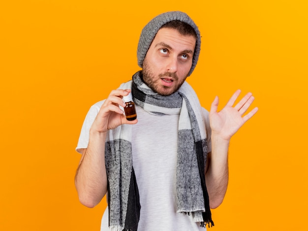 Looking at side unpleased young ill man wearing winter hat and scarf holding medicine in glass bottle and spread hand isolated on yellow background