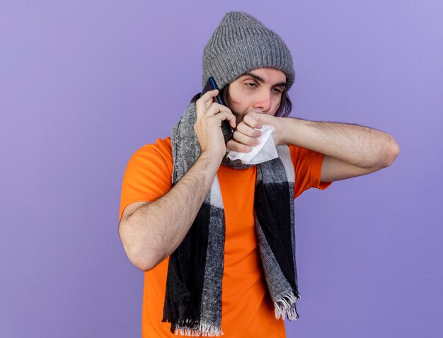 Looking at side weak young ill man wearing winter hat with scarf speaks on phone holding napkin and wiping nose with hand isolated on purple background
