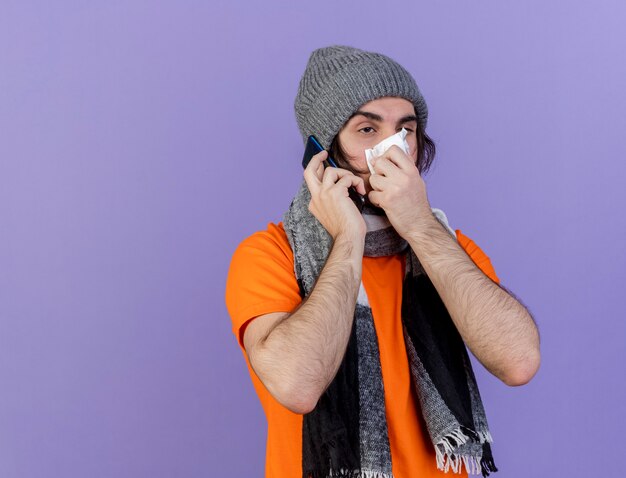 Looking at side weak young ill man wearing winter hat with scarf speaks on phone wiping nose with napkin isolated on purple background