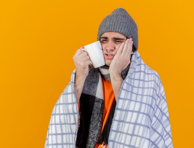 Foto gratuita guardando lato debole giovane uomo malato che indossa il cappello invernale con sciarpa avvolta in mano mettendo plaid