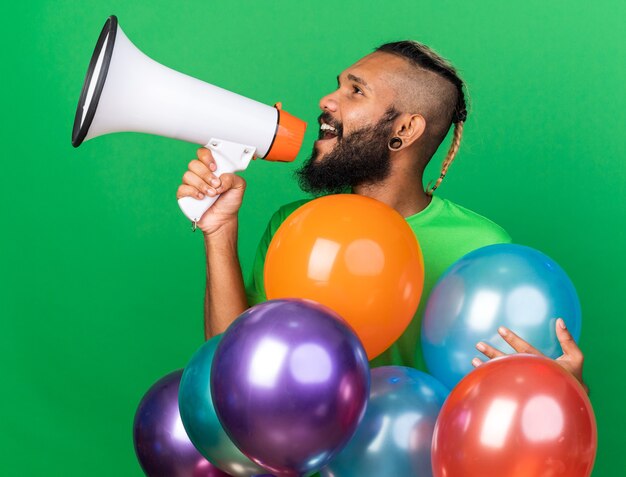 Looking side young afro-american guy wearing green t-shirt standing behind balloons speaks on loudpeaker isolated on green wall