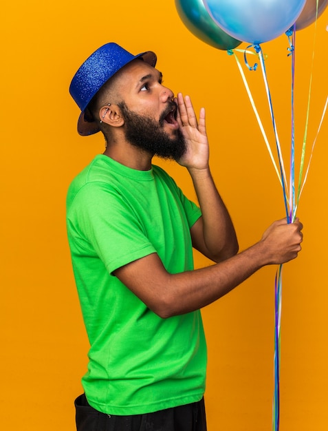 주황색 벽에 고립된 사람을 부르는 풍선을 들고 파티 모자를 쓴 젊은 아프리카계 미국인 남자