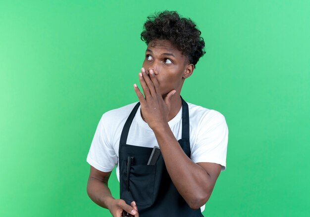 Looking at side young afro-american male barber wearing uniform whispers isolated on green background
