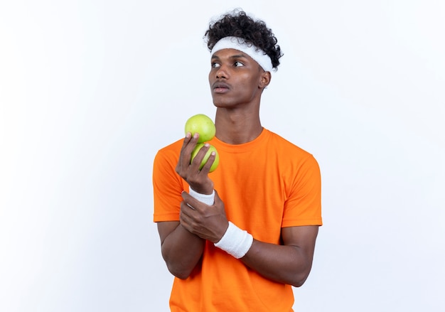 Looking at side young afro-american sporty man wearing headband and wristband holding apples isolated on white background