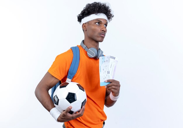 Глядя на сторону молодого афро-американского спортивного человека, носящего повязку на голову и браслет, держа мяч и билеты, изолированные на белом фоне