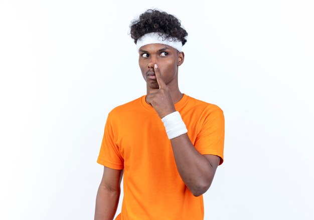 Looking at side young afro-american sporty man wearing headband and wristband putting finger on nose isolated on white background