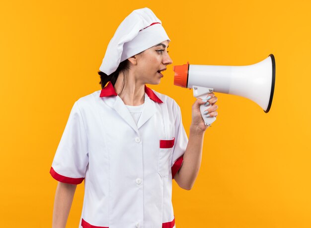 Looking at side young beautiful girl in chef uniform speaks on loudspeaker isolated on orange wall