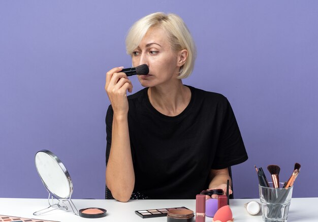 Looking at side young beautiful girl sits at table with makeup tools applying powder blush with powder brush isolated on blue wall