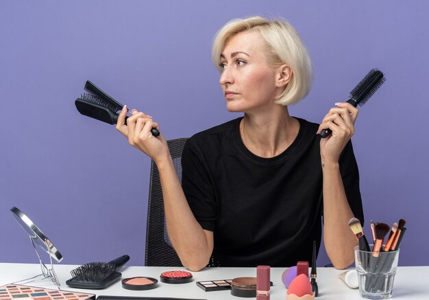 Looking side young beautiful girl sits at table with makeup tools holding combs isolated on blue wall