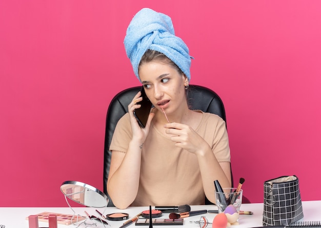 Молодая красивая девушка сидит за столом с инструментами для макияжа, завернув волосы в полотенце, применяя блеск для губ, говорит по телефону, изолированному на розовой стене
