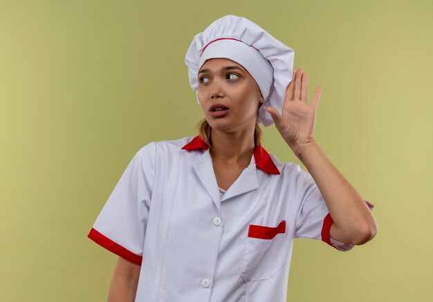 Глядя на сторону молодой женщины-повара в униформе шеф-повара, показывающей жест прослушивания на изолированной зеленой стене с копией пространства