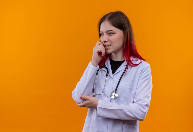 Looking at side young doctor girl wearing stethoscope medical robe putting hand on chin on isolated orange background with copy space