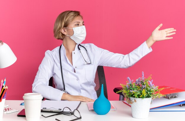 청진 기 및 마스크와 의료 가운을 입고 측면 젊은 여성 의사를 찾고 분홍색 벽에 고립 된 측면에서 손으로 의료 도구 포인트와 책상에 앉아