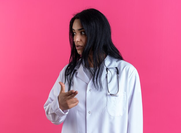 청진 기 분홍색 벽에 고립 된 권총 제스처를 보여주는 의료 가운을 입고 측면 젊은 여성 의사를 찾고