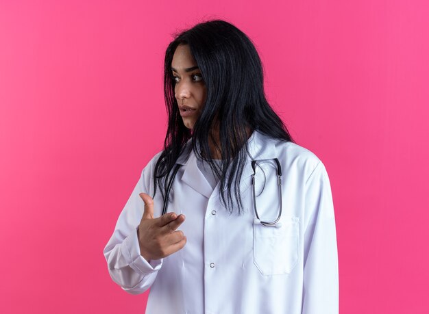 Looking side young female doctor wearing medical robe with stethoscope showing pistol gesture isolated on pink wall